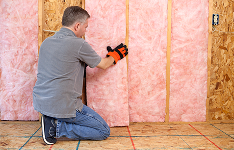 Technician kneeling on floor in unfinished home, installing pink fiberglass batt insulation in a wall.