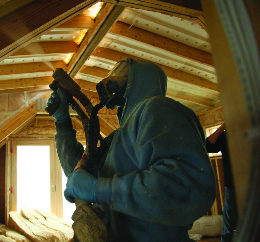 Technician in a hazmat suit, spraying foam insulation into an unfinished attic ceiling.